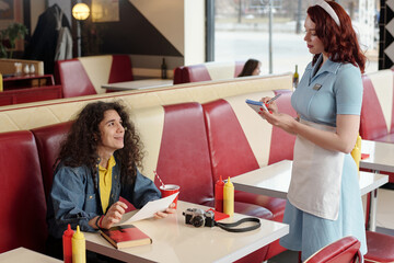 Smiling man with curly hair sitting at diner booth, interacting with waitress writing order on notepad, vintage camera and a notebook on table, ketchup and mustard dispensers - Powered by Adobe