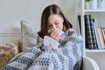 Young sick woman sitting under warm blanket sneezing coughing in handkerchief
