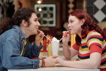 Young couple sharing milkshake in retro-themed diner, both enjoying their time together. Blurred colorful background enhances nostalgic feel, creating warm and cozy atmosphere
