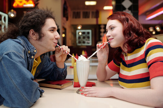 Young couple sitting in retro diner sharing milkshake while smiling and looking at each other without a care in the world enjoying the nostalgic setting, adding to the romantic atmosphere