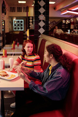 Two young women sharing conversation and food in vintage diner booth with milkshakes, fries, and burger around them under vibrant lighting in the background