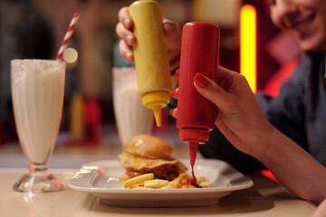 Close-up of someone's hands squeezing mustard and ketchup onto fries with milkshake and burger in...