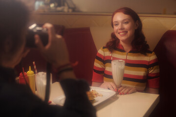 Portrait of young woman smiling while sitting in retro diner booth, red-striped sweater and milkshake on table, photographer capturing moment