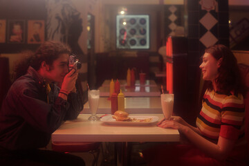 Capturing candid photograph in retro-themed diner setting with friends. One person holding vintage camera, another person enjoying milkshake and burger