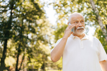 Naklejka premium Handsome gray-haired senior man with white beard smiling talking on smartphone in sunny park, enjoying fresh air and greenery. Confident elderly man with white beard enjoying leisurely phone call.