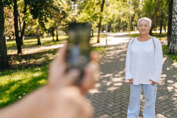 Selective focus shot of gray-haired senior woman posing happily in sunny park, while husband photographer capturing moment with smartphone, lush green background adding to serene scene.