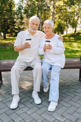 Fototapeta premium Full length vertical portrait of loving senior couple enjoying coffee sitting hugging on park bench, basking in summer warmth, exuding love and happiness, embodying love and happiness in retirement.