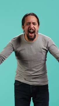 Vertical video Portrait of irate man feeling outraged, yelling at friend, releasing pent up emotion, studio backdrop. Furious person full of rage screaming at mate isolated over blue studio background