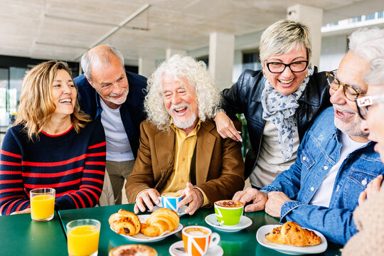 Happy group of senior people laughing and enjoying coffee at cafeteria bar. Retired generation community having fun gathered on terrace toasting hot drink. Elderly friendship lifestyle concept.