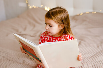 The girl in festive pajamas is lying on the bed and reading a big book