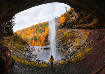 Female Hiker in Yellow Coat Adventure Travel Autumn New England Waterfall Cave. Kaaterskill Falls with Peak Fall Foliage Colorful Forest of Trees