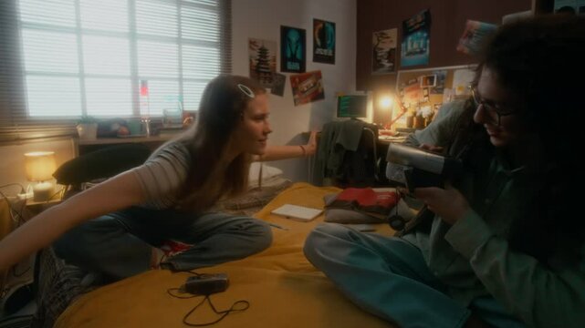 Handheld full shot of smiling puffy haired boy recording cheerful gen Y girl who waving hands and dancing to retro vhs camera while hanging out together in bedroom in 90s
