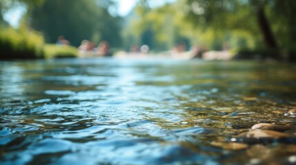 Fototapeta premium A tranquil shallow stream with clear water flows through a scenic park on a sunny day. Trees and blurred figures in the background contribute to a peaceful, outdoor atmosphere.