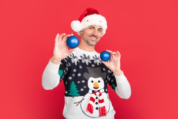 Studio portrait of guy wears santa hat and winter sweater. Santa with bauble, christmas ball. Man with christmas hat over isolated red background.