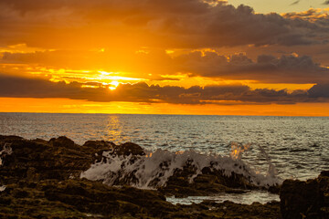 Dramatic sunset over the ocean with waves crashing against the rocky shore. The intense orange and yellow colors of the sky contrast with the dark rocks and foaming waves.