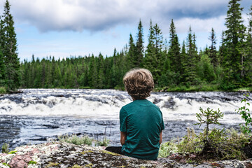 A young blond boy sitting on rocky riverbank, gazing at the rushing rapids of Storån River, surrounded by a dense forest on a warm summer day in the Nordic Wilderness