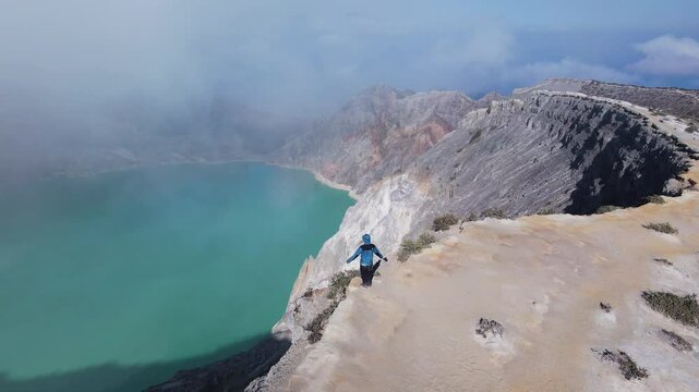 Man standing at the top of a mountain with a view of turquoise sulfur water lake, surrounded by rock cliff at Kawah Ijen volcano, East Java, Indonesia.