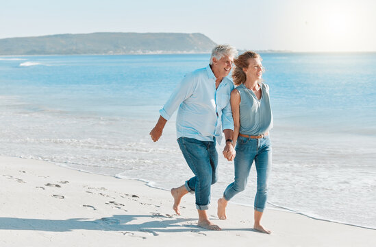 Mature couple, running and holding hands with beach for holiday, weekend or outdoor bonding. Man, woman or lovers with smile for vacation, stroll or view in sunshine on sandy shore by ocean coast