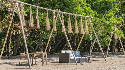 Fototapeta premium A relaxation area on a tropical beach. Wicker plastic furniture: tables, chairs, a sofa with soft cushions stand on the sand. Decorative tassels are suspended on bamboo poles. Lush green vegetation.