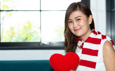 Portrait young woman smiling good looking black hair, lady holding red heart looking at camera with positive expression, wearing checkered shirt. Indoor studio shot winter windows background.