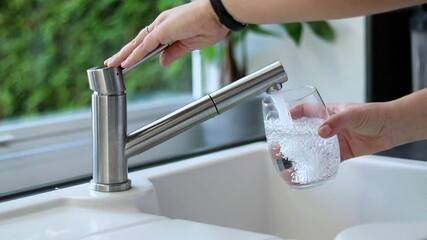Close up of female hands pouring tap water into a glass in the kitchen. White sink and blurred background 