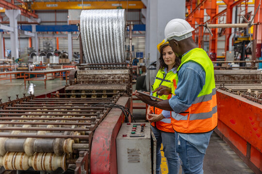 Two mechanical engineers from different cultures working together to check a steel making production system by using a tablet. Cooperation between workers in a steel factory. Workers in safety suit