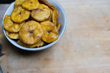 Keripik pisang or banana crackers on a white bowl at wooden cutting board. With copy space. Indonesian traditional snack made from banana and sugar.