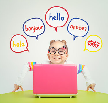Foreign language learning concept of a beautiful cute little girl with eyeglasses behind a laptop on table and looking at camera. Hello word in Spanish, French, Russian and Chinese. 