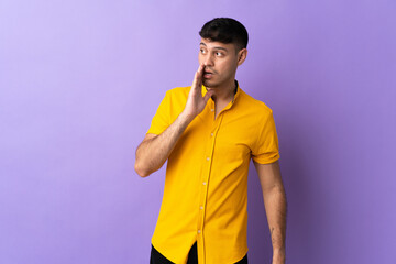 Young Colombian man isolated on purple background whispering something with surprise gesture while...