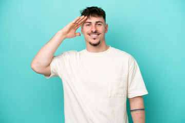 Young caucasian handsome man isolated on blue background saluting with hand with happy expression