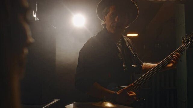 Medium shot of Caucasian male musician wearing hat playing electric guitar in company of unrecognizable female bandmate playing percussions during jam session at recording studio