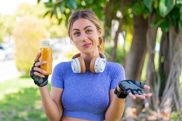 Young pretty sport woman holding an orange juice at outdoors making doubts gesture while lifting...
