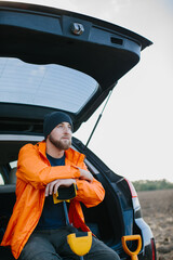 A male treasure hunter sits in the trunk of his car leaning on a metal detector.