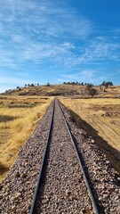 Captures a set of railway tracks stretching into the distance through a dry, grassy landscape. The clear sky and the horizon marked by low hills create a sense of vastness and journey.
