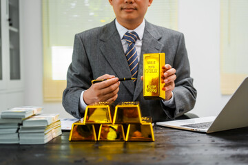 Businessman in suit sits at his desk, holding a shining gold bar. financial charts, the scene...