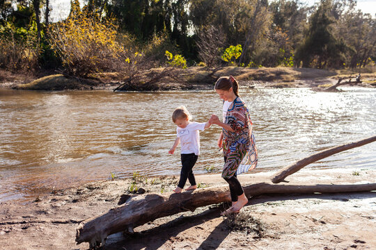 Big sister holding hand of little brother as he balances on log by river