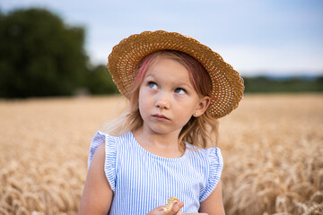 Child girl in a hat stands against the background of a wheat field