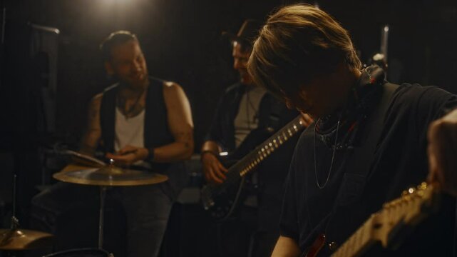 Medium shot of group of young Caucasian male musicians strumming electric guitars and having friendly conversation while prepaing for life performance