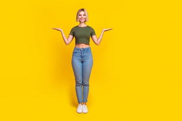 Full size photo of cheerful girl dressed khaki t-shirt jeans palms comparing products empty space isolated on yellow color background