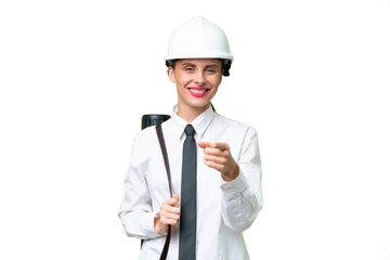 Young architect woman with helmet and holding blueprints over isolated background surprised and pointing front