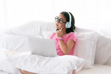  Asian young girl wearing glasses and headphones sitting on white bed using laptop for study online, learning or entertainment. Relaxed indoor environment with focus on education and technology.