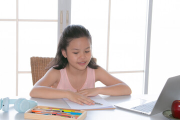 Obraz premium Adorable Asian girl online learning, writing notes while using a laptop, elementary school girl typing answer or response with teacher, Focus on digital communication and learning.