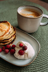 plate of homemade syrniki with whipped cream and cranberries, served with a cup of coffee on green woven placemat in cozy breakfast setting