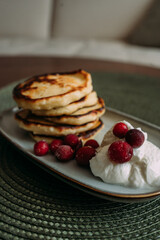 stack of homemade pancakes with whipped cream and fresh cranberries on ceramic plate, placed on green woven placemat in cozy kitchen setting