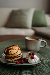 plate of homemade syrniki with whipped cream and cranberries, served with a cup of coffee on green woven placemat in cozy breakfast setting