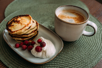 plate of homemade syrniki with whipped cream and cranberries, served with a cup of coffee on green woven placemat in cozy breakfast setting