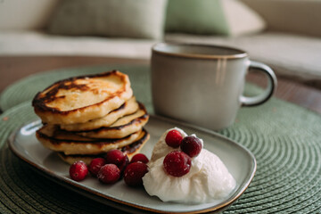 plate of homemade syrniki with whipped cream and cranberries, served with a cup of coffee on green woven placemat in cozy breakfast setting