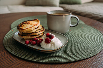 plate of homemade syrniki with whipped cream and cranberries, served with a cup of coffee on green woven placemat in cozy breakfast setting