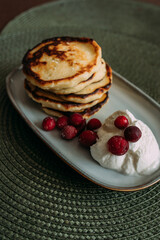 stack of homemade pancakes with whipped cream and fresh cranberries on ceramic plate, placed on green woven placemat in cozy kitchen setting