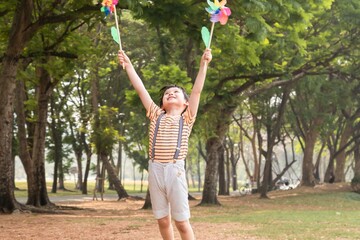 Obraz premium Young Asian boy holding two colorful pinwheels in both hands, smiling happily outdoors in a park setting, wearing striped shirt with suspenders, sunny day surrounded by green trees, joyful expression